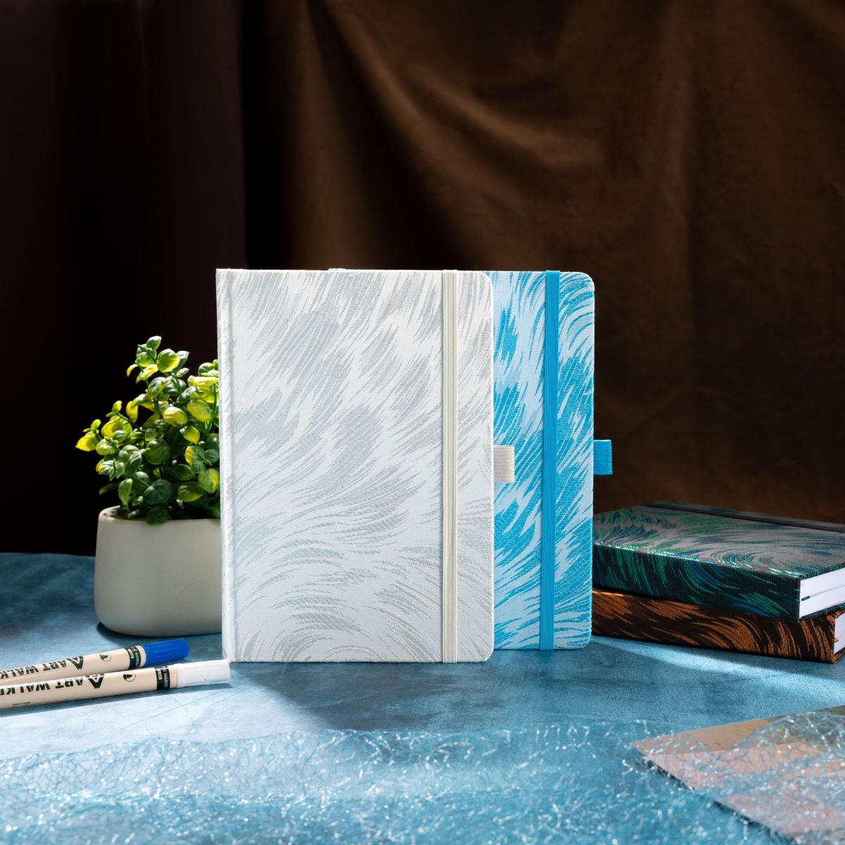 `Two A5 hardcover notebooks displayed upright against a dark draped background with stationery accessories`