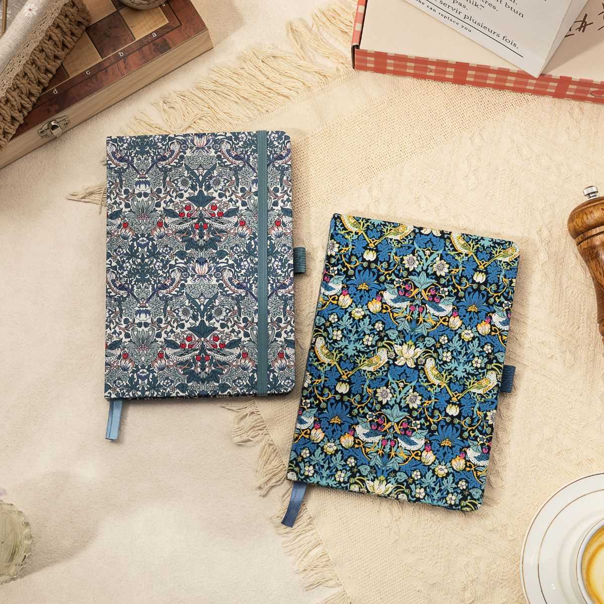 A5 floral fabric notebook with a blue bird and botanical pattern, shown alongside a matching notebook on a soft neutral textile surface.