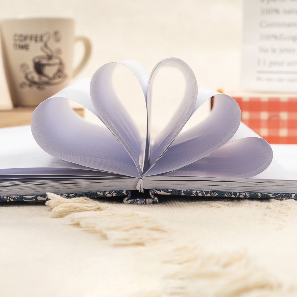 An open journal with pages folded into a heart shape, resting on a table with a "COFFEE TIME" mug in the background, capturing a warm and cozy writing moment.