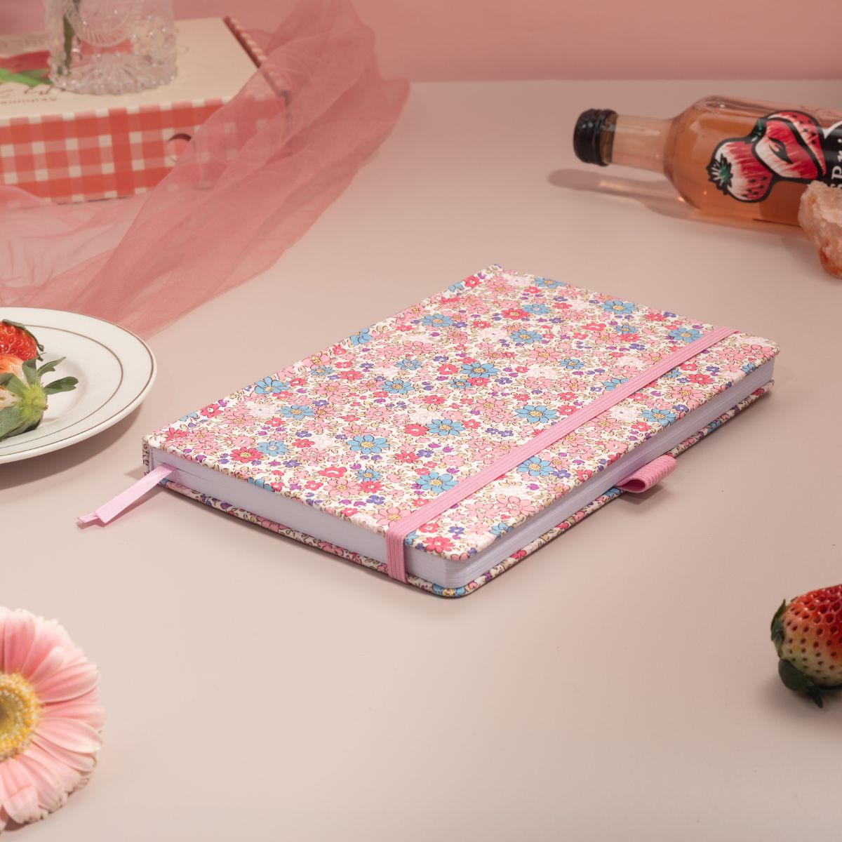A pink woven fabric notebook with a delicate floral print, laid flat on a pink table, surrounded by fresh strawberries, a decorative bottle, and pink crystals, evoking a peaceful, screen-free writing ritual.
