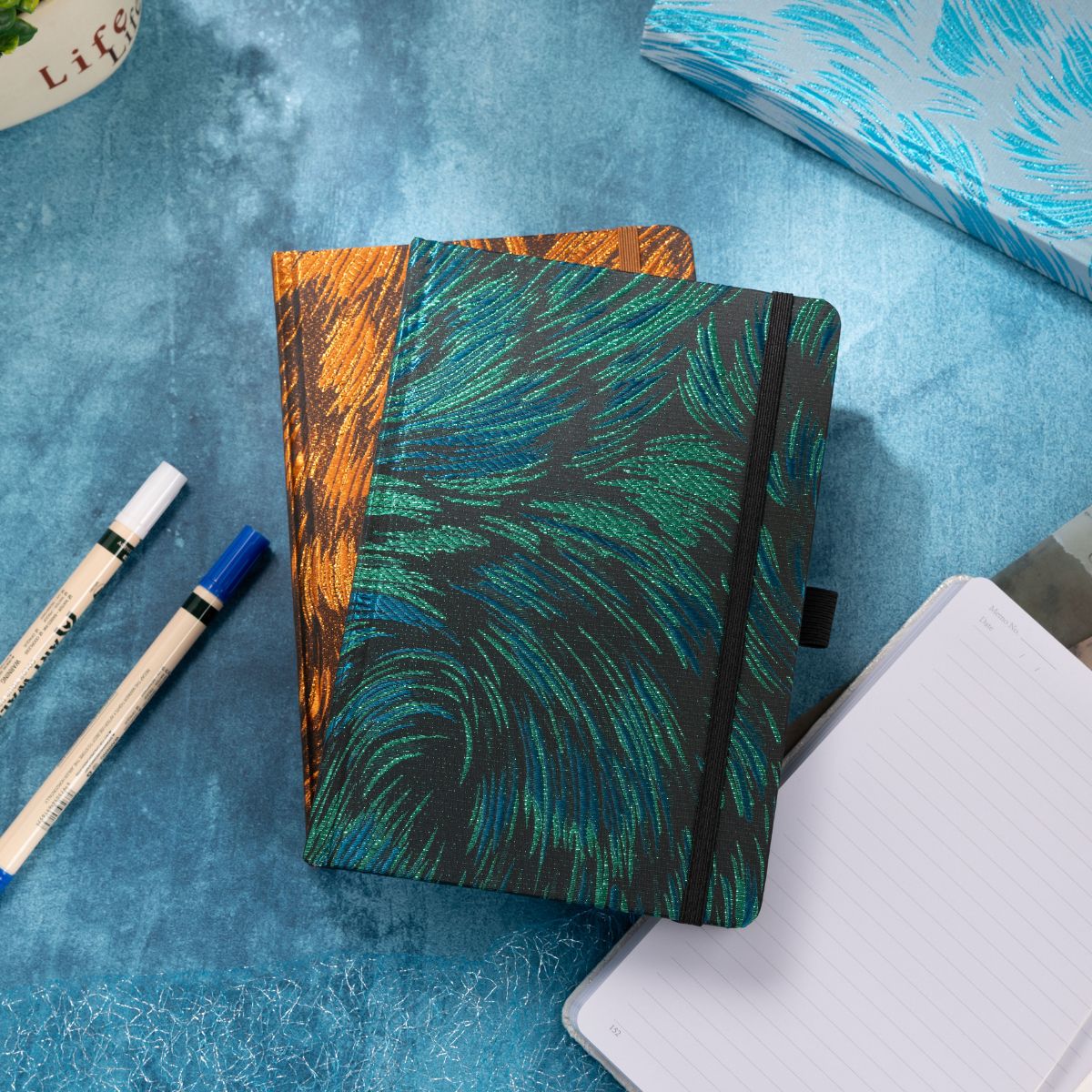 Two embroidered fabric notebooks stacked on a blue textured surface, featuring teal and black feather-like stitching and a copper-brown pattern, styled with white markers and an open journal.