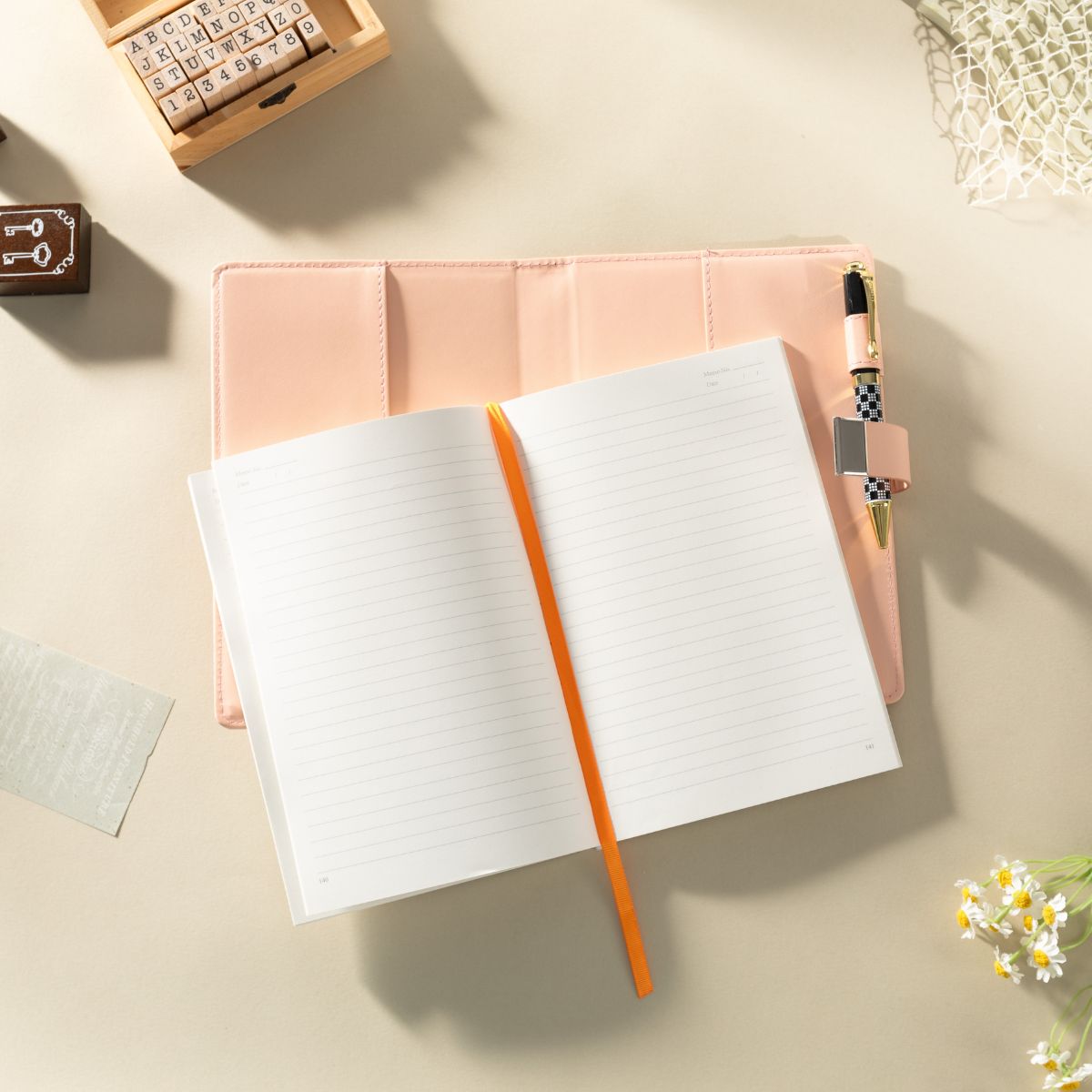 Open lined notebook on a pale pink cover with an orange ribbon bookmark and a pen on a light surface