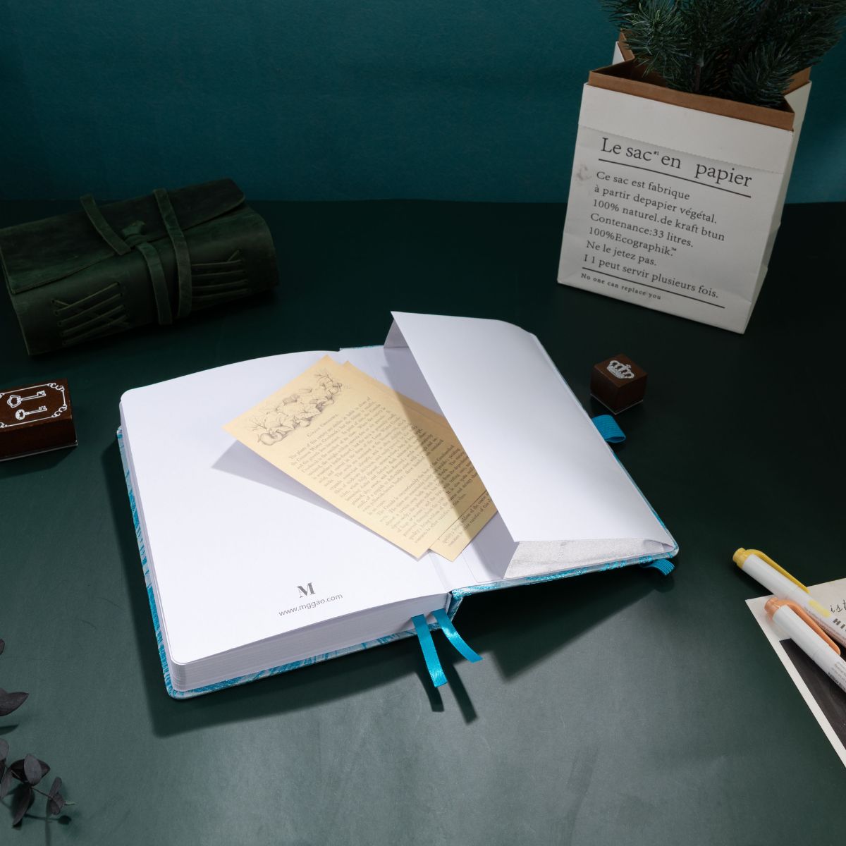 A light blue embroidered A5 journal open on a dark green desk, featuring a blue ribbon bookmark, a vintage card, and a paper bag planter, creating a peaceful and aesthetic writing corner.