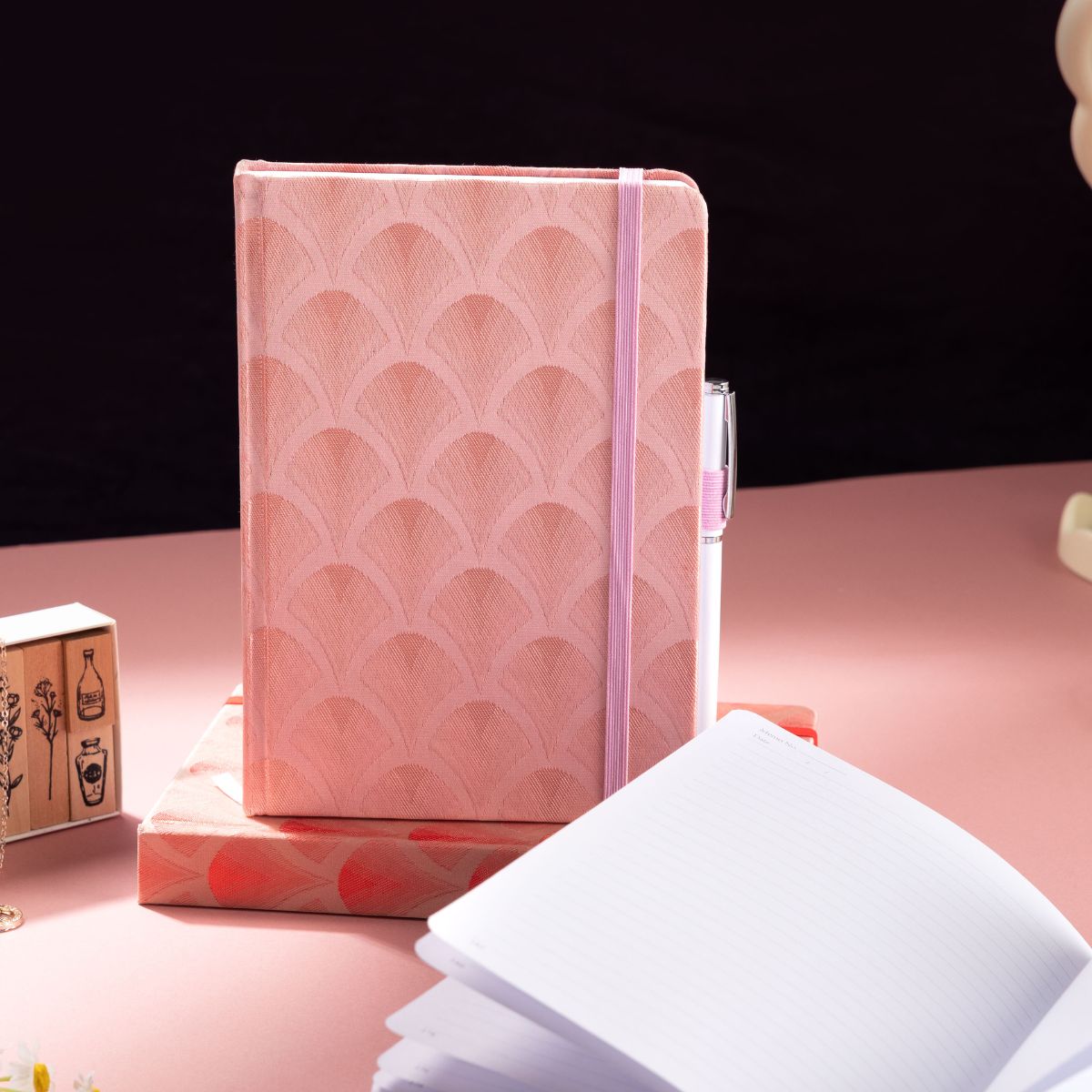 Pink fabric hardcover notebook with a patterned cover, elastic closure, and lined pages on a pink desk.