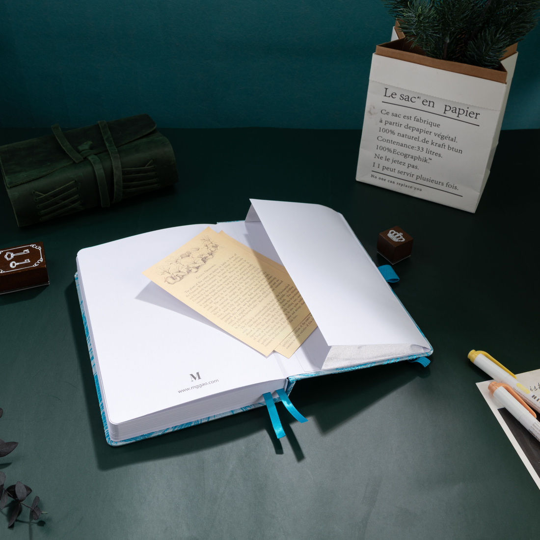 A light blue embroidered A5 journal open on a dark green desk, featuring a blue ribbon bookmark, a vintage card, and a paper bag planter, creating a peaceful and aesthetic writing corner.