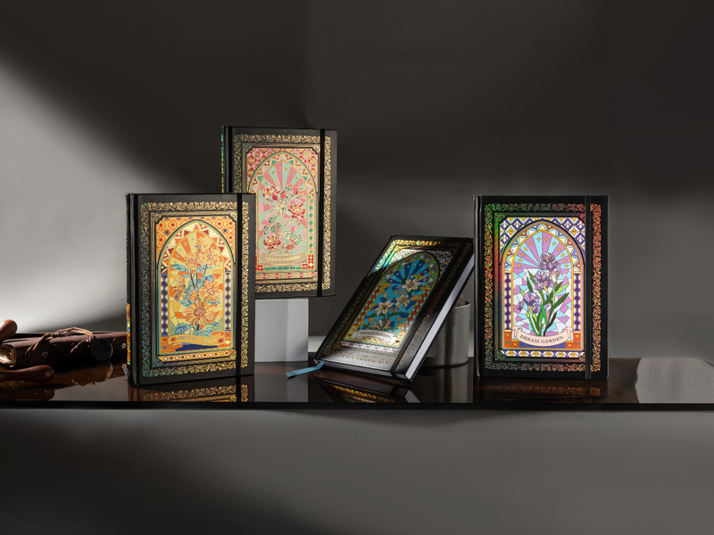 A collection of four Dream Garden holographic cardboard notebooks featuring rose, sunflower, lily, and iris floral arch designs, arranged on a dark reflective desk with a brown leather pencil case, showcasing the full series lineup.