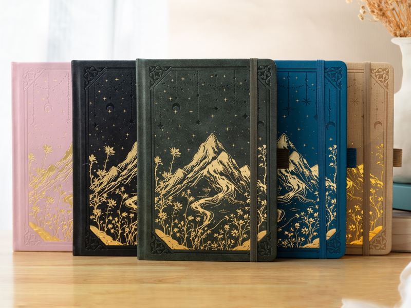 A collection of gilded vegan leather notebooks featuring gold foil mountain and starry night embossing in black, dark green, navy blue, pink, and beige, lined up on a wooden desk.