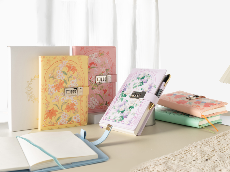 A collection of floral vegan leather lockable notebooks in yellow, pink, purple, and blue, featuring a 3-digit metal combination lock, styled with a white gift box and lace fabric on a light desk.