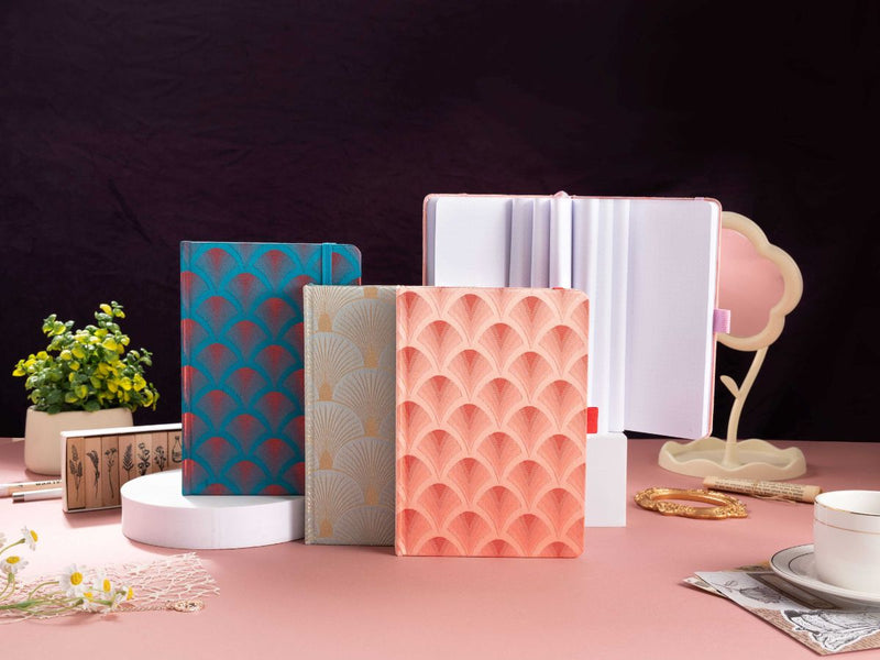 A collection of woven fabric notebooks featuring scalloped embroidery in teal, beige, and peach, displayed on a pink desk with a green plant, wooden stamps, and vintage decor.