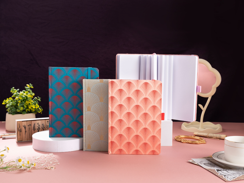 A collection of woven fabric notebooks featuring scalloped embroidery in teal, beige, and peach, displayed on a pink desk with a green plant, wooden stamps, and vintage decor.