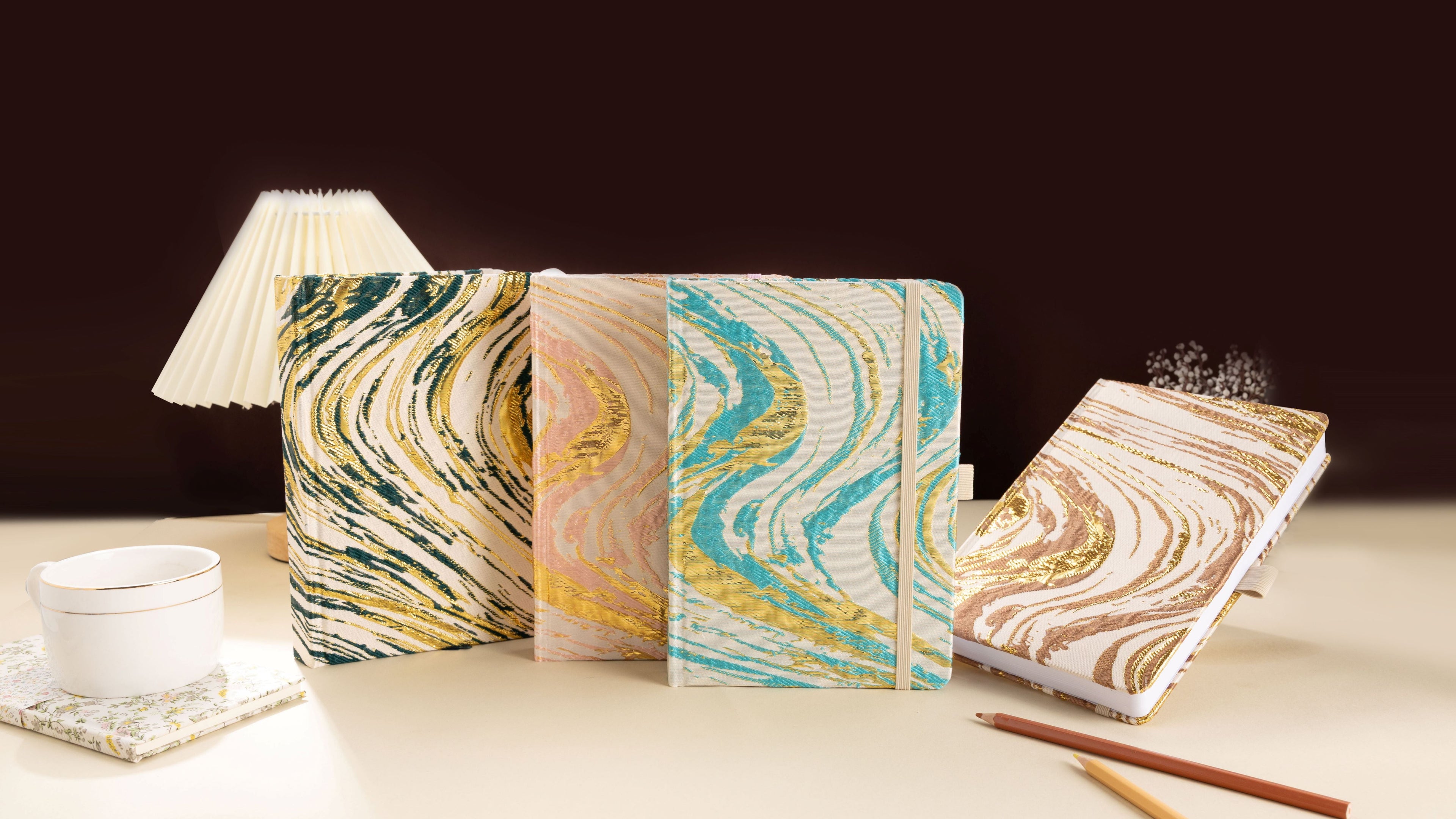 Four A5 woven fabric notebooks with metallic marble swirls in emerald gold, blush pink, teal gold, and bronze brown, arranged on a cream desk with a pleated lamp, coffee cup, and pencils.