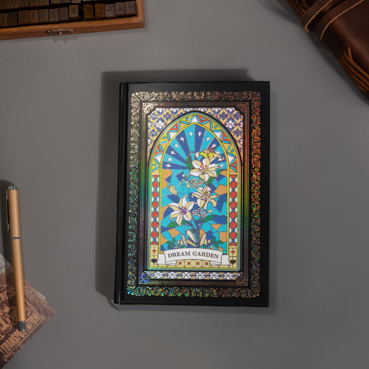 Black and blue holographic lily Dream Garden cardboard notebook lying flat on a gray surface, showcasing the full floral arch design with vibrant iridescent borders and a black elastic closure, styled with a wooden stamp box and a gold pen.