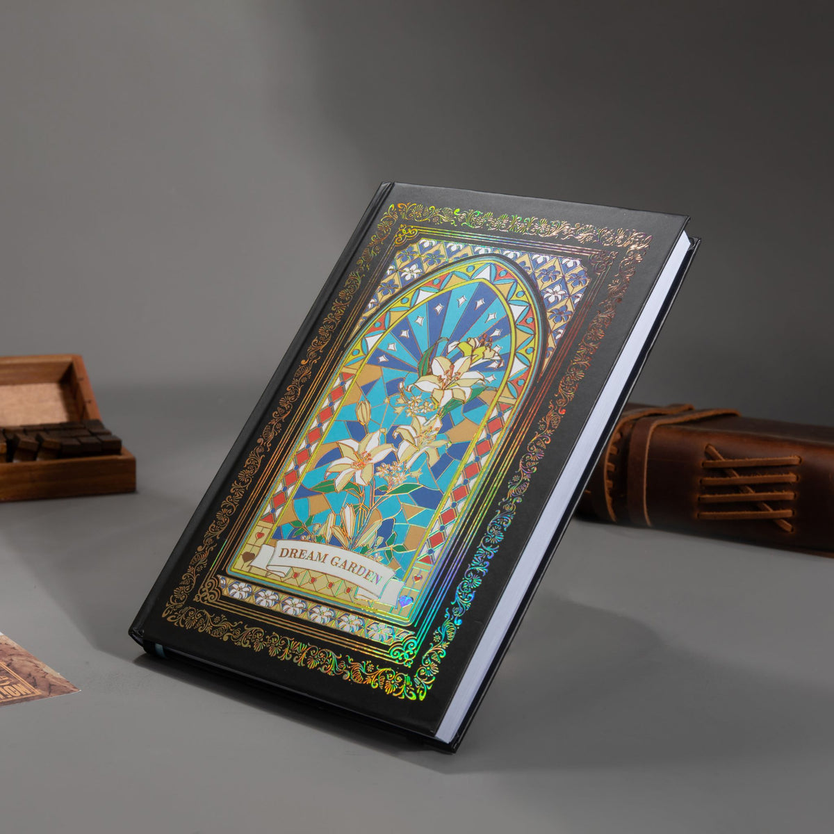 Black and blue holographic lily Dream Garden cardboard notebook resting angled on a gray surface, featuring a white lily floral arch design with iridescent borders, styled with wooden stamps and a brown leather notebook.