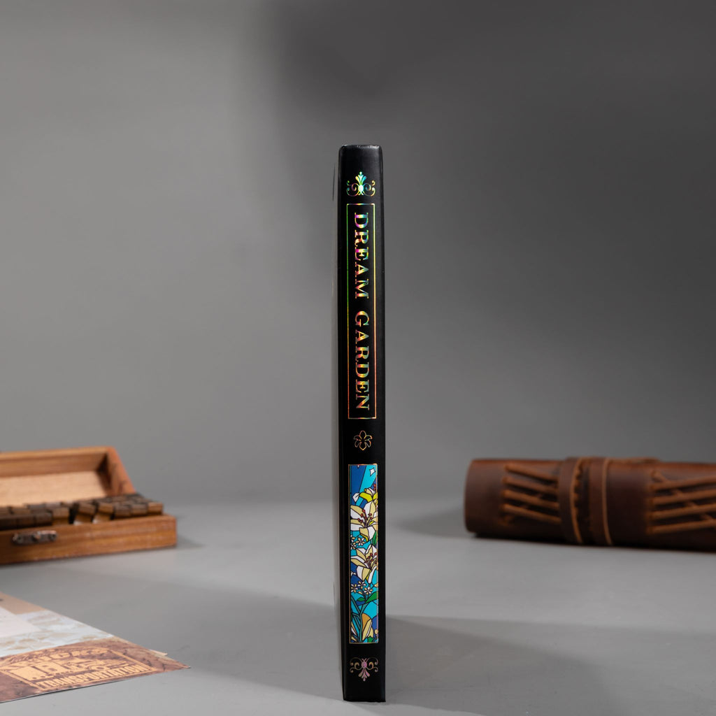 Spine view of the black lily Dream Garden cardboard notebook standing upright on a dark surface, featuring the holographic "DREAM GARDEN" title and geometric patterns.