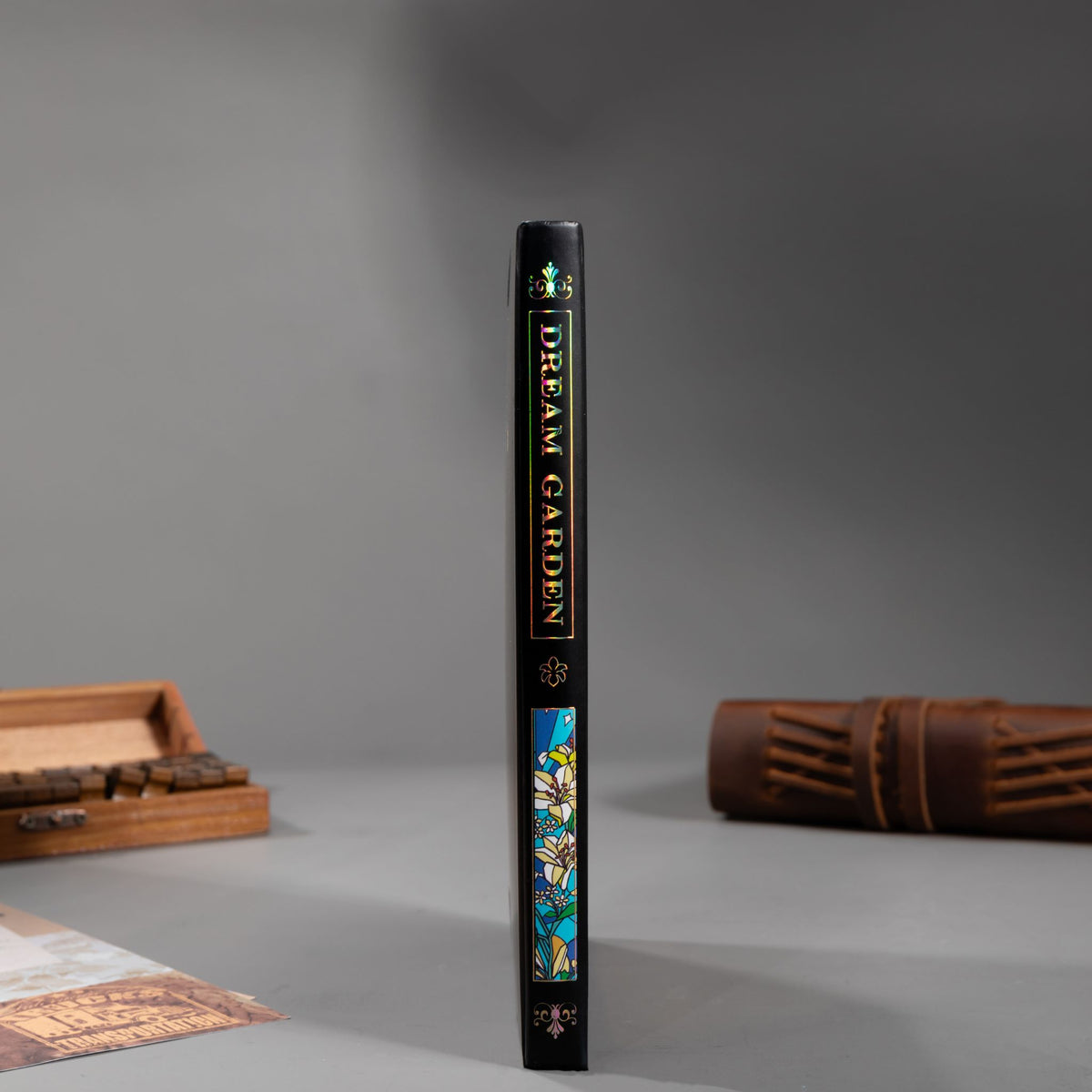 Spine view of the black lily Dream Garden cardboard notebook standing upright on a dark surface, featuring the holographic "DREAM GARDEN" title and geometric patterns.