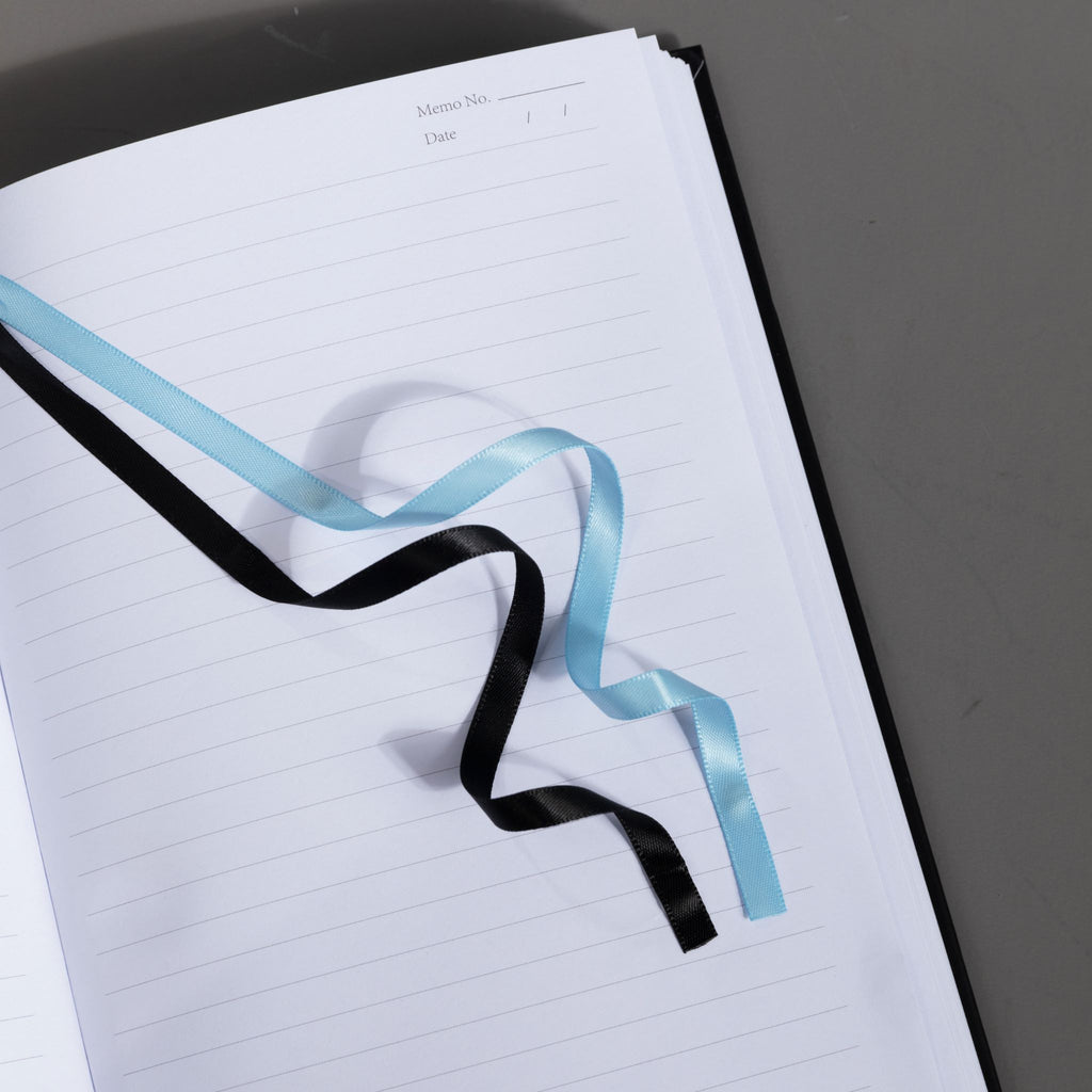 Close-up of the black and blue two-tone ribbon bookmark resting elegantly on the open lined pages of the lily Dream Garden notebook.