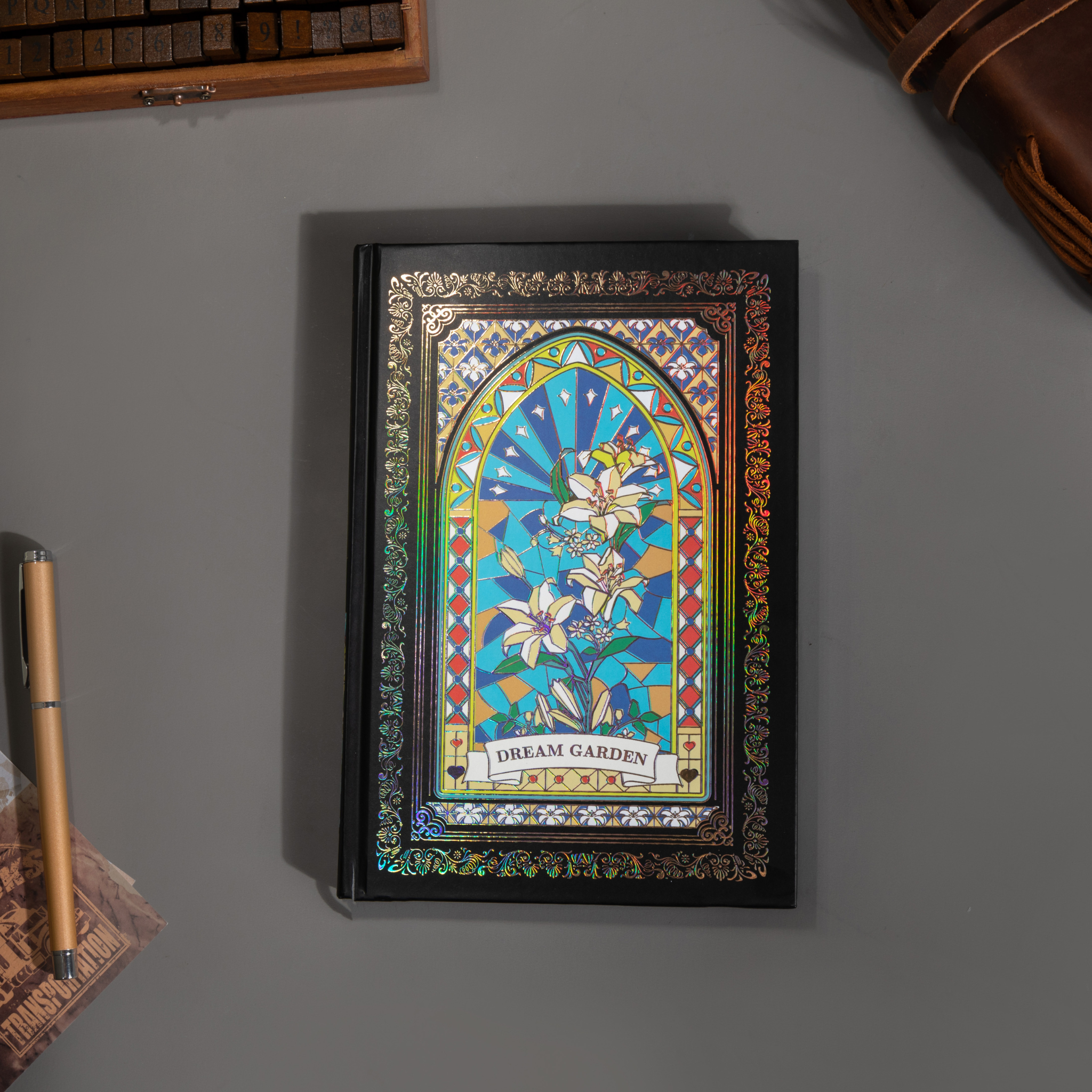 Black and blue holographic lily Dream Garden cardboard notebook lying flat on a gray surface, showcasing the full floral arch design with vibrant iridescent borders and a black elastic closure, styled with a wooden stamp box and a gold pen.
