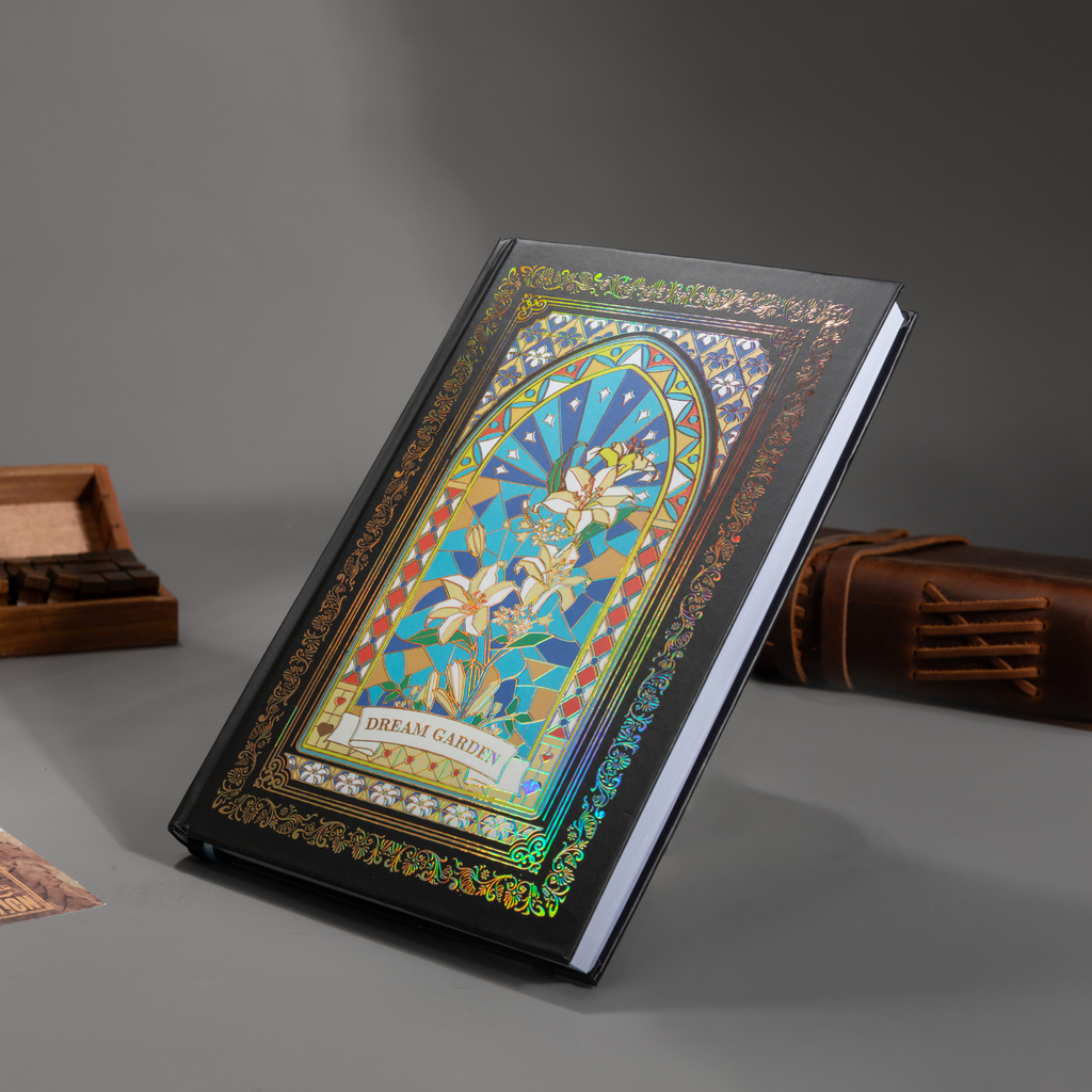 Black and blue holographic lily Dream Garden cardboard notebook resting angled on a gray surface, featuring a white lily floral arch design with iridescent borders, styled with wooden stamps and a brown leather notebook.