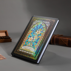 Black and blue holographic lily Dream Garden cardboard notebook resting angled on a gray surface, featuring a white lily floral arch design with iridescent borders, styled with wooden stamps and a brown leather notebook.