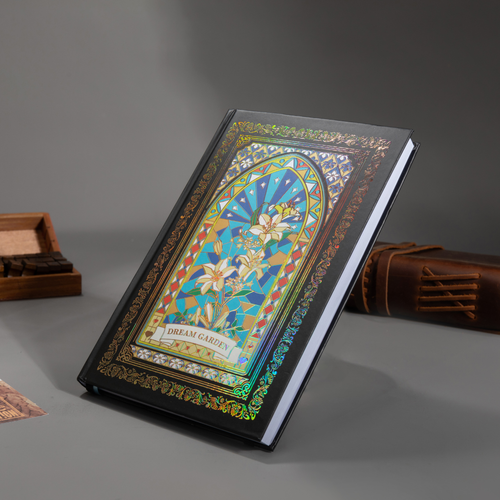 Black and blue holographic lily Dream Garden cardboard notebook resting angled on a gray surface, featuring a white lily floral arch design with iridescent borders, styled with wooden stamps and a brown leather notebook.