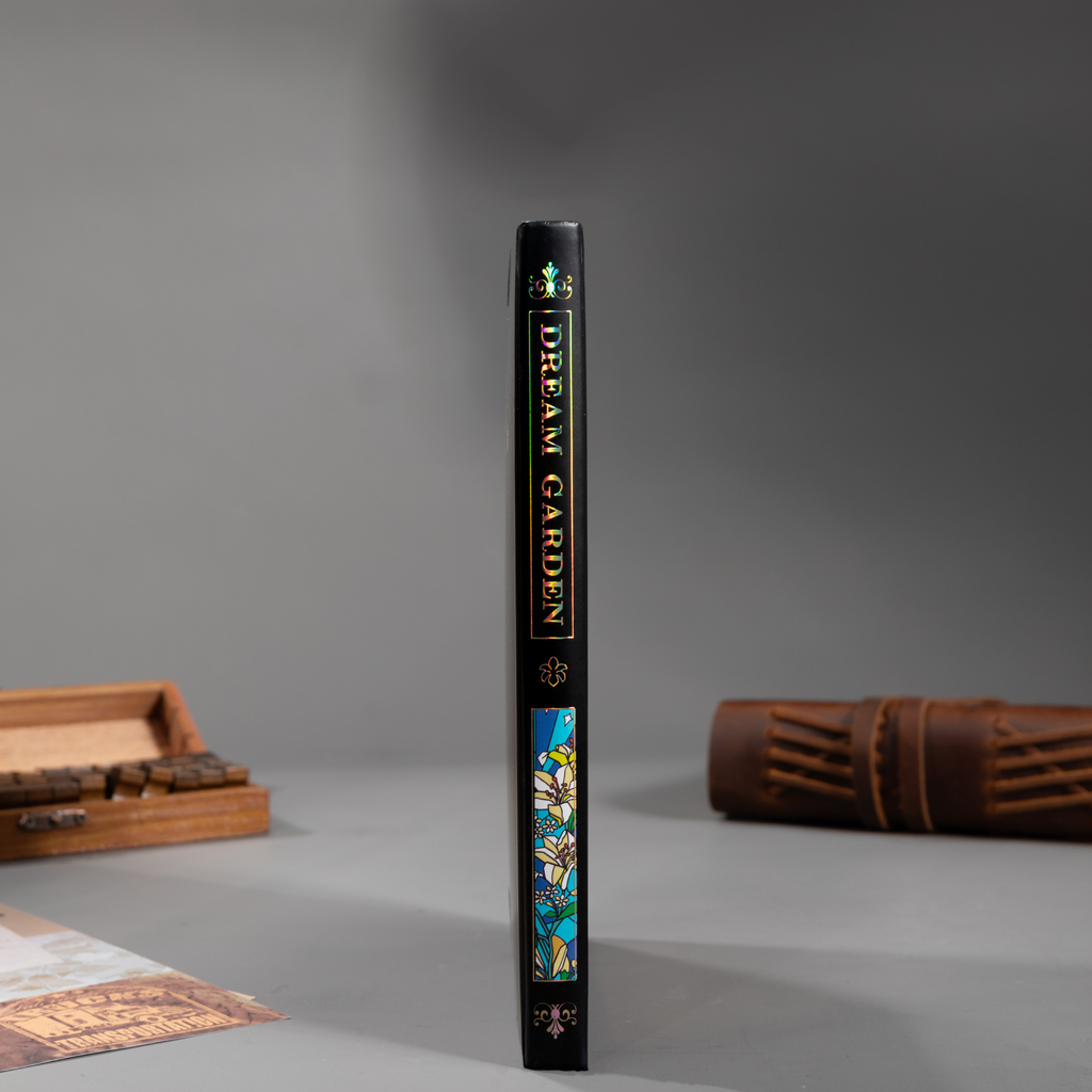 Spine view of the black lily Dream Garden cardboard notebook standing upright on a dark surface, featuring the holographic "DREAM GARDEN" title and geometric patterns.