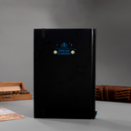 Back cover of the black lily Dream Garden cardboard notebook standing upright, featuring a minimalist "DREAM GARDEN" logo and star design, styled with wooden stamps and books on a dark desk.