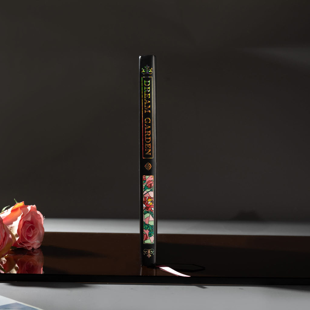 Spine view of the black Dream Garden cardboard notebook standing upright on a dark reflective surface, featuring the holographic title "DREAM GARDEN" and floral details.
