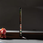 Spine view of the black Dream Garden cardboard notebook standing upright on a dark reflective surface, featuring the holographic title "DREAM GARDEN" and floral details.
