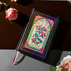 Angled flat lay of the black and pink holographic Dream Garden cardboard notebook resting on a dark reflective surface, featuring a vibrant floral arch design with holographic neon accents, and styled with fresh pink roses.