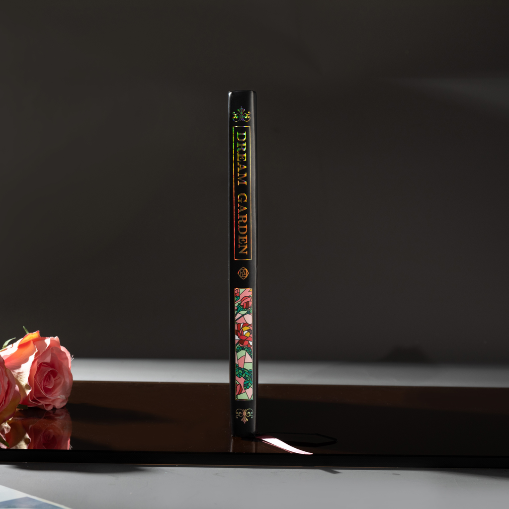Spine view of the black Dream Garden cardboard notebook standing upright on a dark reflective surface, featuring the holographic title "DREAM GARDEN" and floral details.