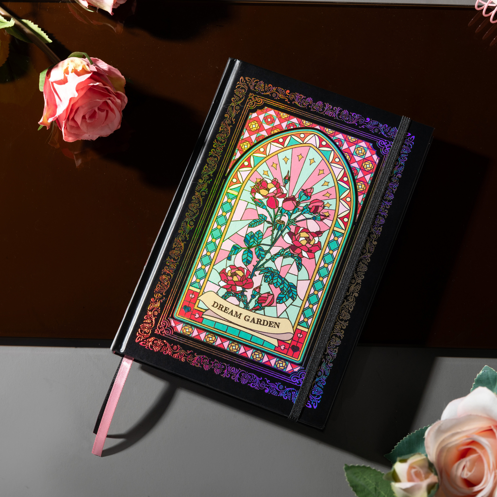 Angled flat lay of the black and pink holographic Dream Garden cardboard notebook resting on a dark reflective surface, featuring a vibrant floral arch design with holographic neon accents, and styled with fresh pink roses.