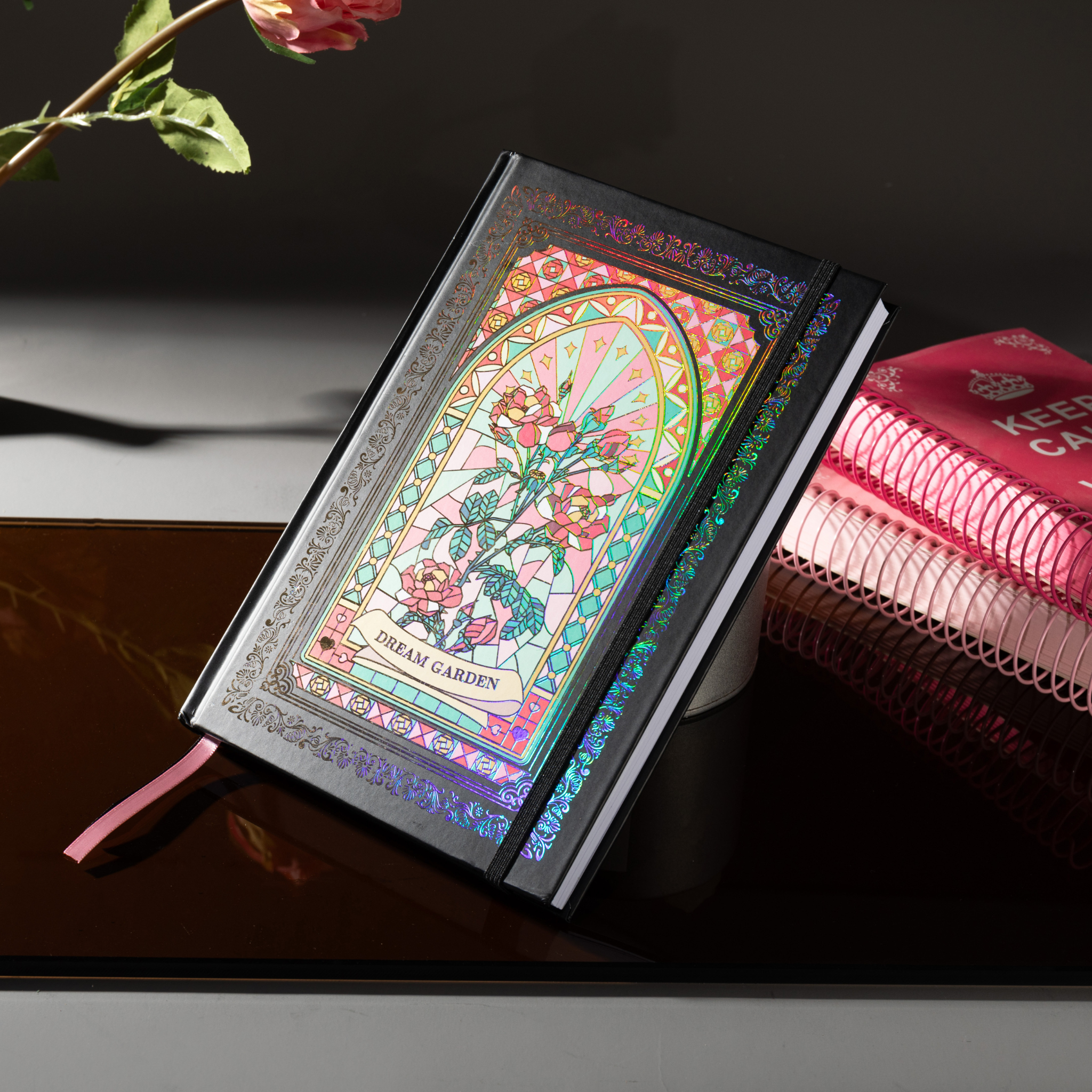 Black and pink holographic Dream Garden cardboard notebook resting angled on a dark reflective surface, styled with pink roses.