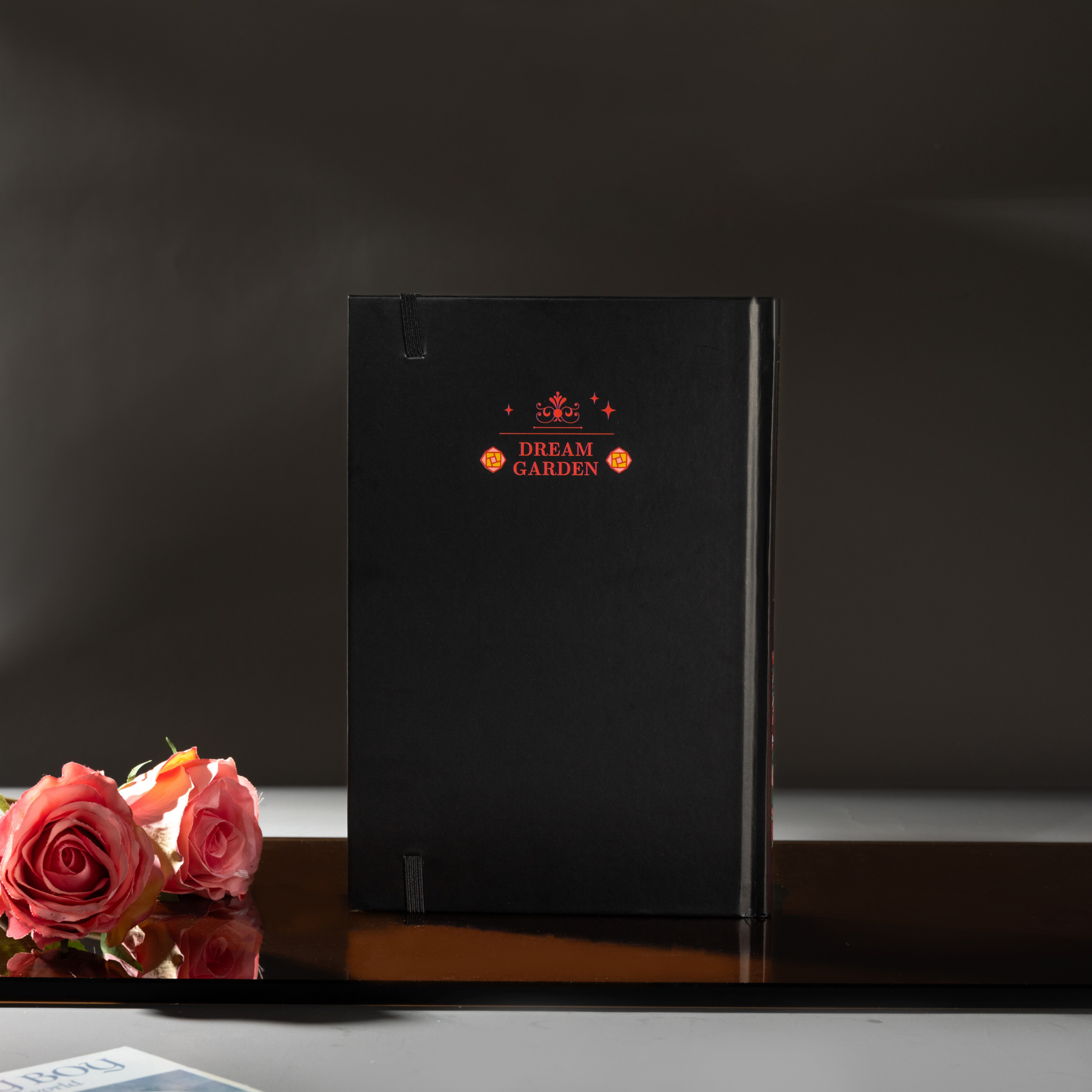 Back cover of the black Dream Garden cardboard notebook standing upright, featuring a minimalist "DREAM GARDEN" logo and star design, styled with pink roses.