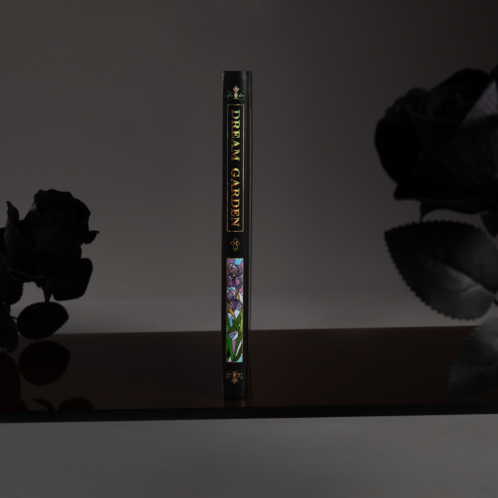 Spine view of the black iris Dream Garden cardboard notebook standing upright on a dark surface, featuring the holographic "DREAM GARDEN" title and geometric details.