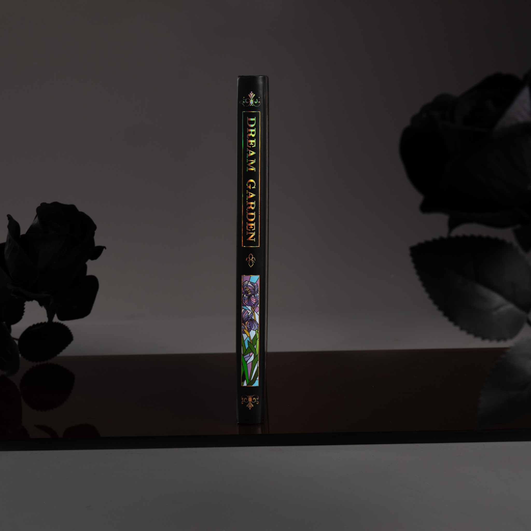 Spine view of the black iris Dream Garden cardboard notebook standing upright on a dark surface, featuring the holographic "DREAM GARDEN" title and geometric details.