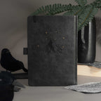Elegant back cover of the black vegan leather notebook featuring embossed gold foil stars and moon, styled on a moody desk setup.