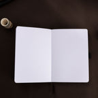 Open A5 journal laid flat on a dark surface, displaying the clean 160gsm dot grid paper interior and a black ribbon bookmark.