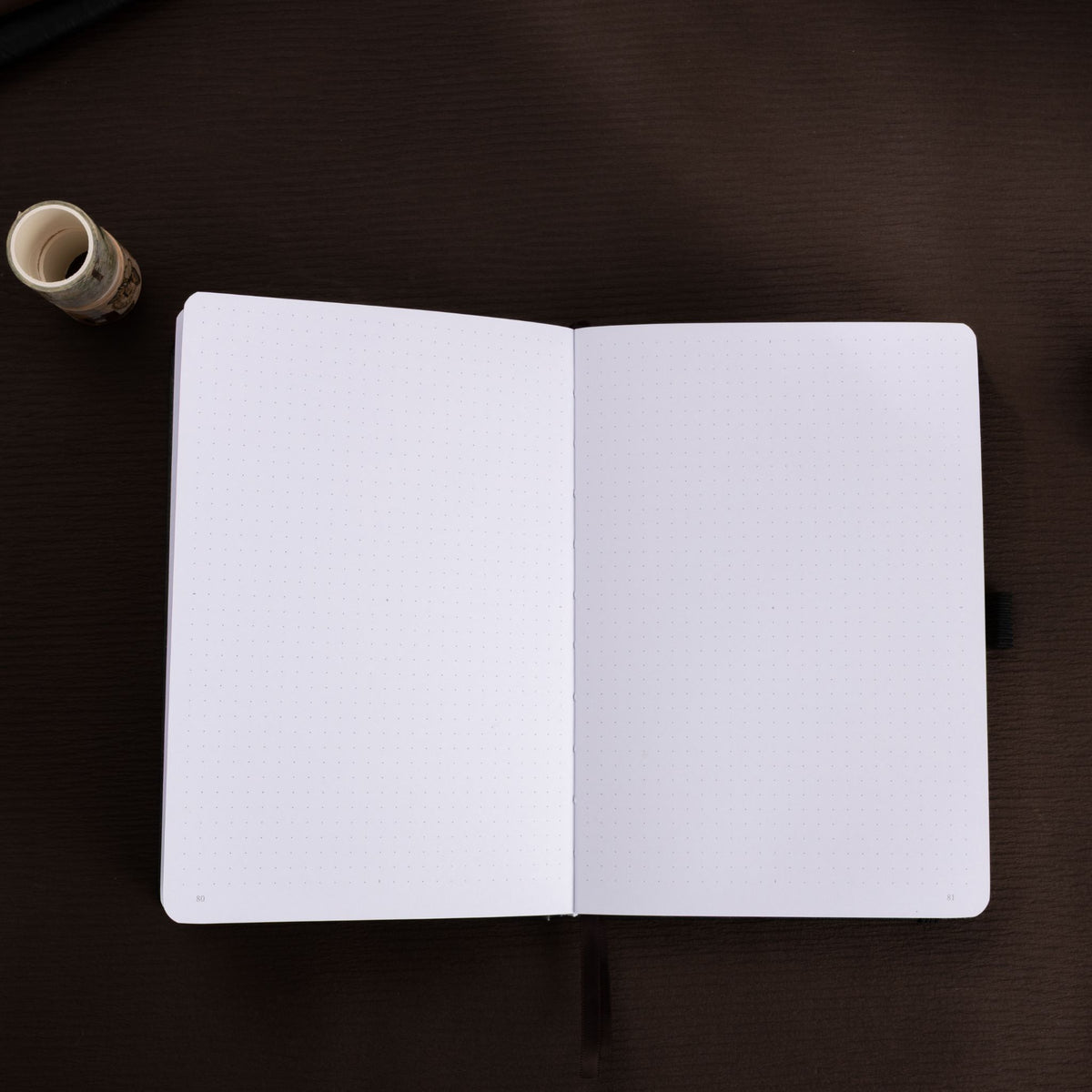 Open A5 journal laid flat on a dark surface, displaying the clean 160gsm dot grid paper interior and a black ribbon bookmark.