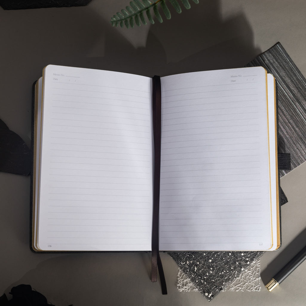 Open A5 notebook lying flat, showing the 100gsm lined paper interior with a black ribbon bookmark and black side pen loop.