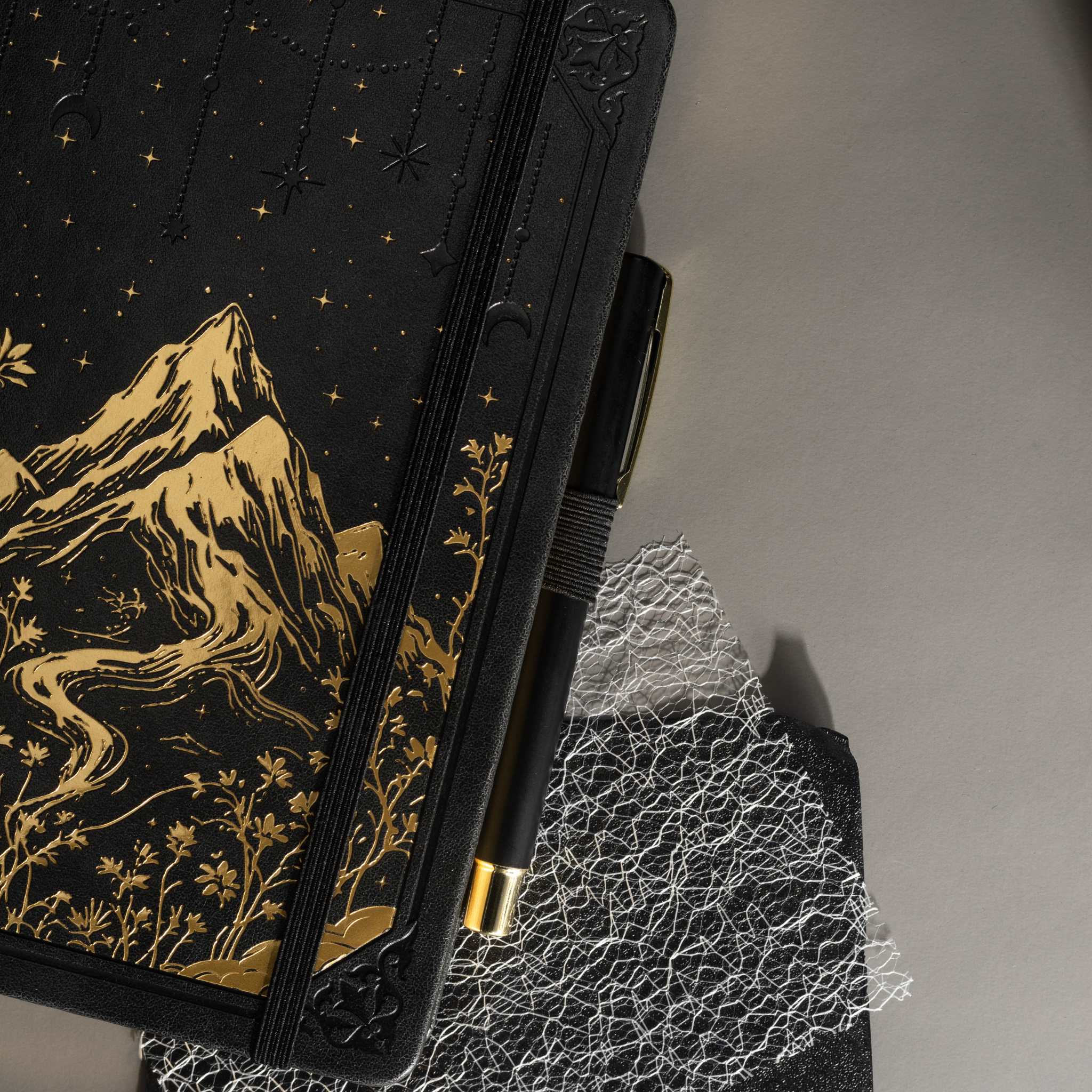 Angled view of the black A5 mountain notebook resting on a dark surface, showing the gold foil cover details and elastic band.