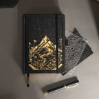 Black vegan leather journal with gold mountain embossing laid flat on a desk with a matching black pen and dark accessories.