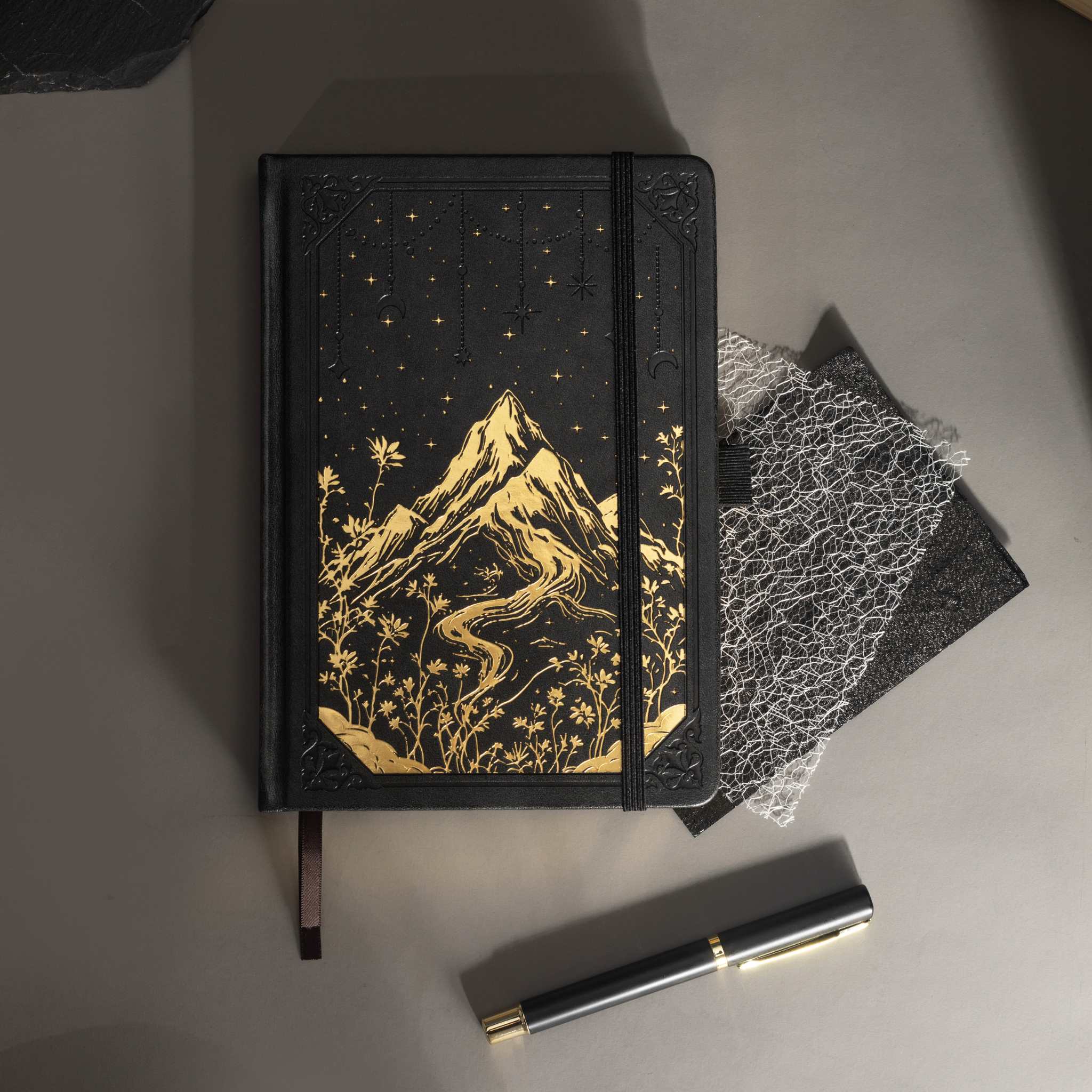Black vegan leather journal with gold mountain embossing laid flat on a desk with a matching black pen and dark accessories.