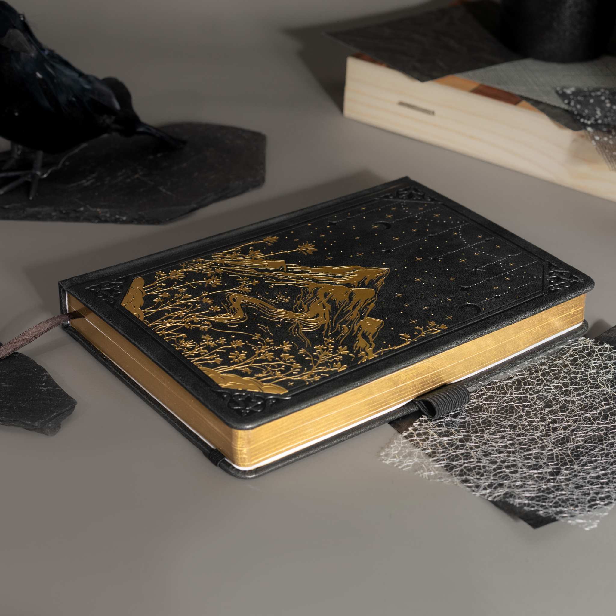 Black vegan leather notebook lying flat on a gray surface, showcasing the gold foil mountain and starry night cover design, gold gilded edges, a black ribbon bookmark, and a black elastic band.