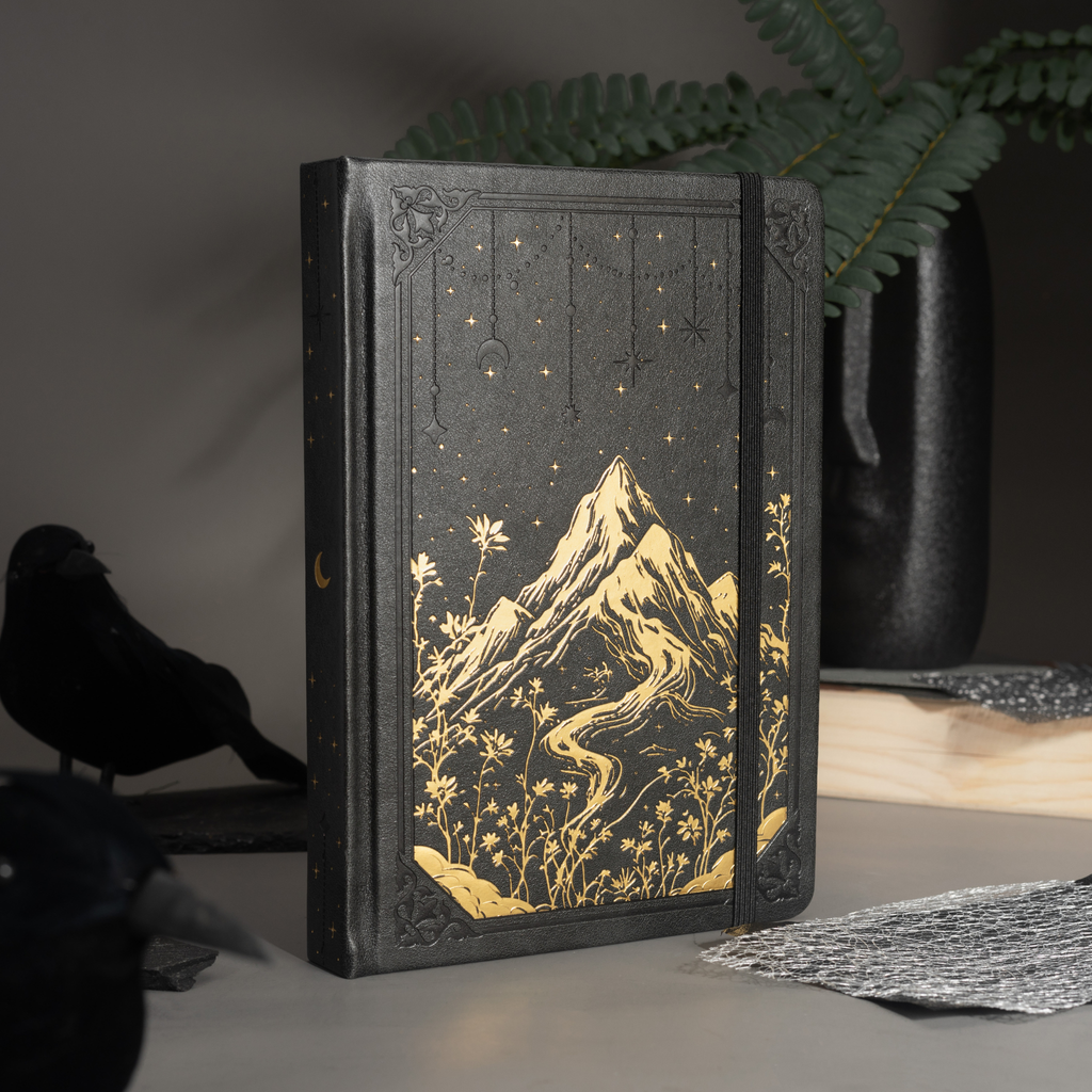 Black vegan leather notebook standing upright on a dark wooden desk with a black pen, showcasing the gold foil mountain cover.