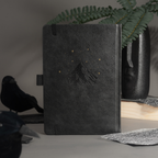 Elegant back cover of the black vegan leather notebook featuring embossed gold foil stars and moon, styled on a moody desk setup.