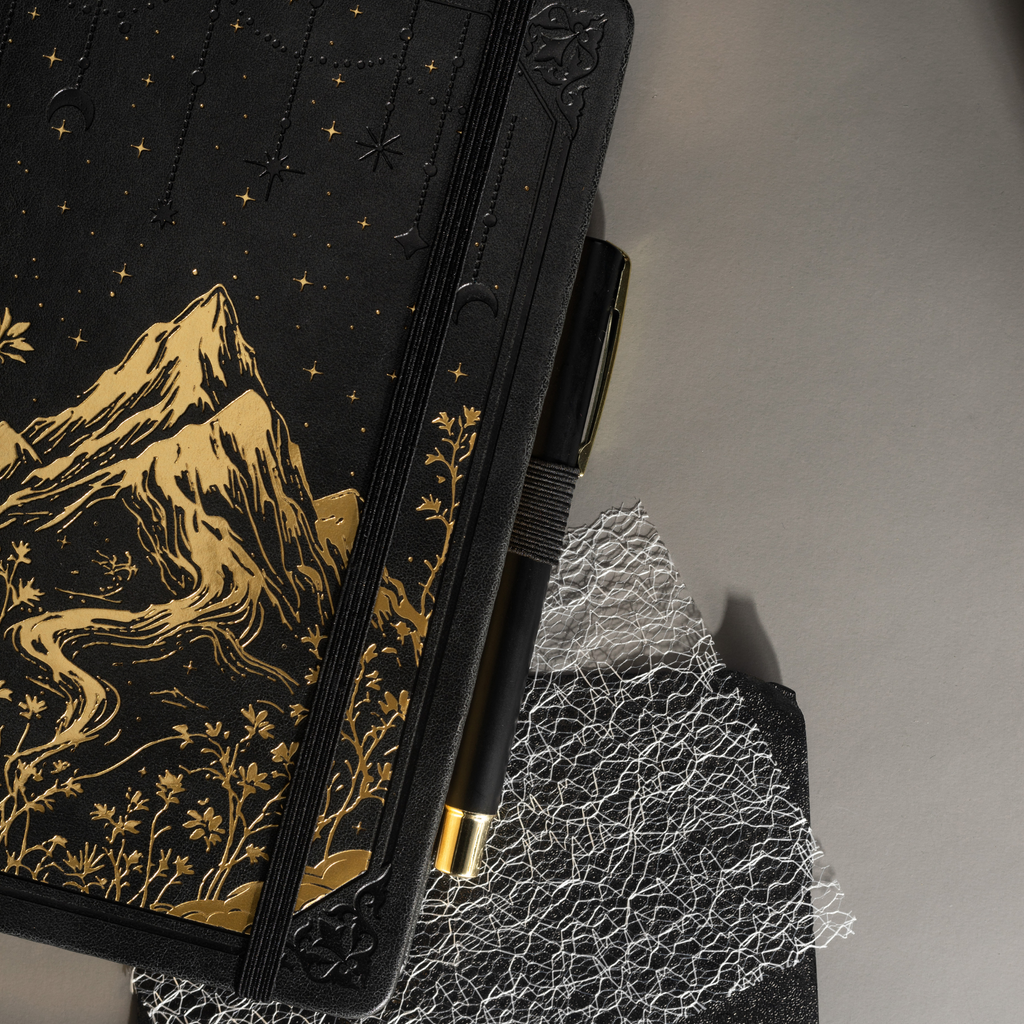 Angled view of the black A5 mountain notebook resting on a dark surface, showing the gold foil cover details and elastic band.