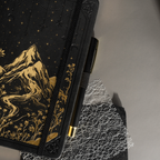 Angled view of the black A5 mountain notebook resting on a dark surface, showing the gold foil cover details and elastic band.