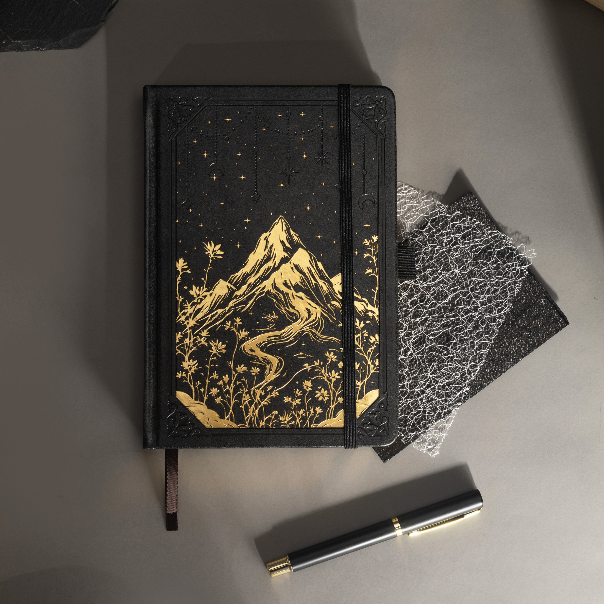 Black vegan leather journal with gold mountain embossing laid flat on a desk with a matching black pen and dark accessories.