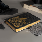 Black vegan leather notebook lying flat on a gray surface, showcasing the gold foil mountain and starry night cover design, gold gilded edges, a black ribbon bookmark, and a black elastic band.