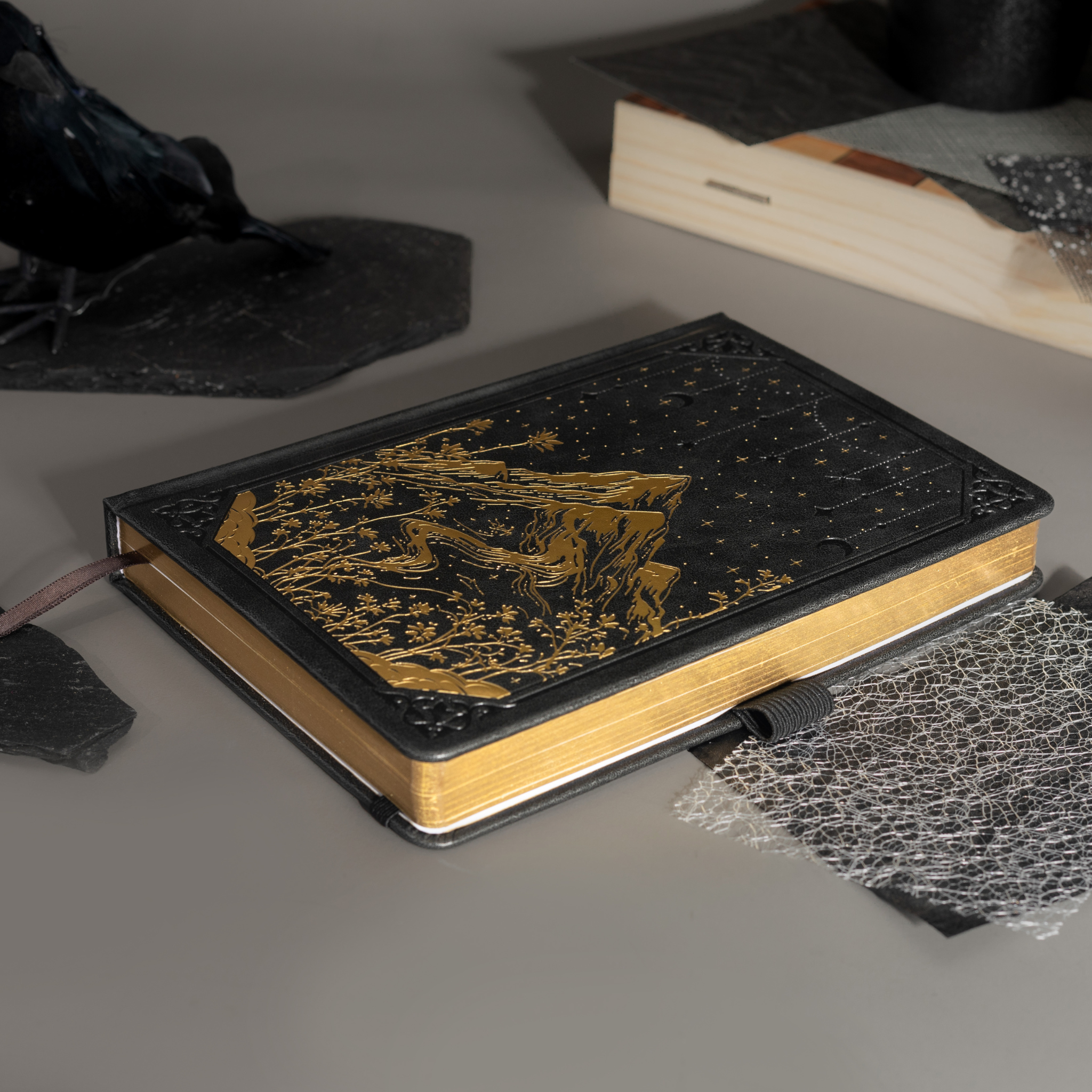 Black vegan leather notebook lying flat on a gray surface, showcasing the gold foil mountain and starry night cover design, gold gilded edges, a black ribbon bookmark, and a black elastic band.